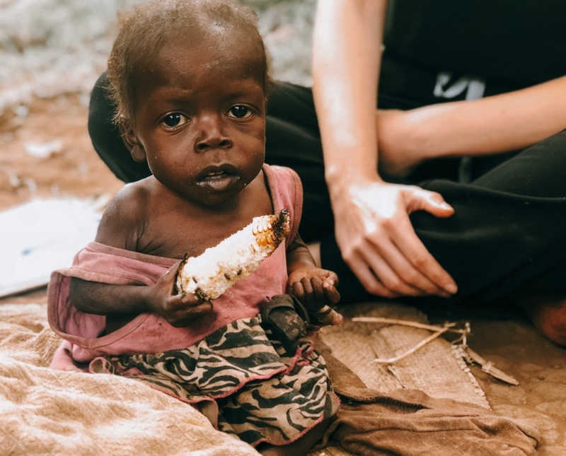 Woman sits on ground of orphanage beside sick little girl holding bread
