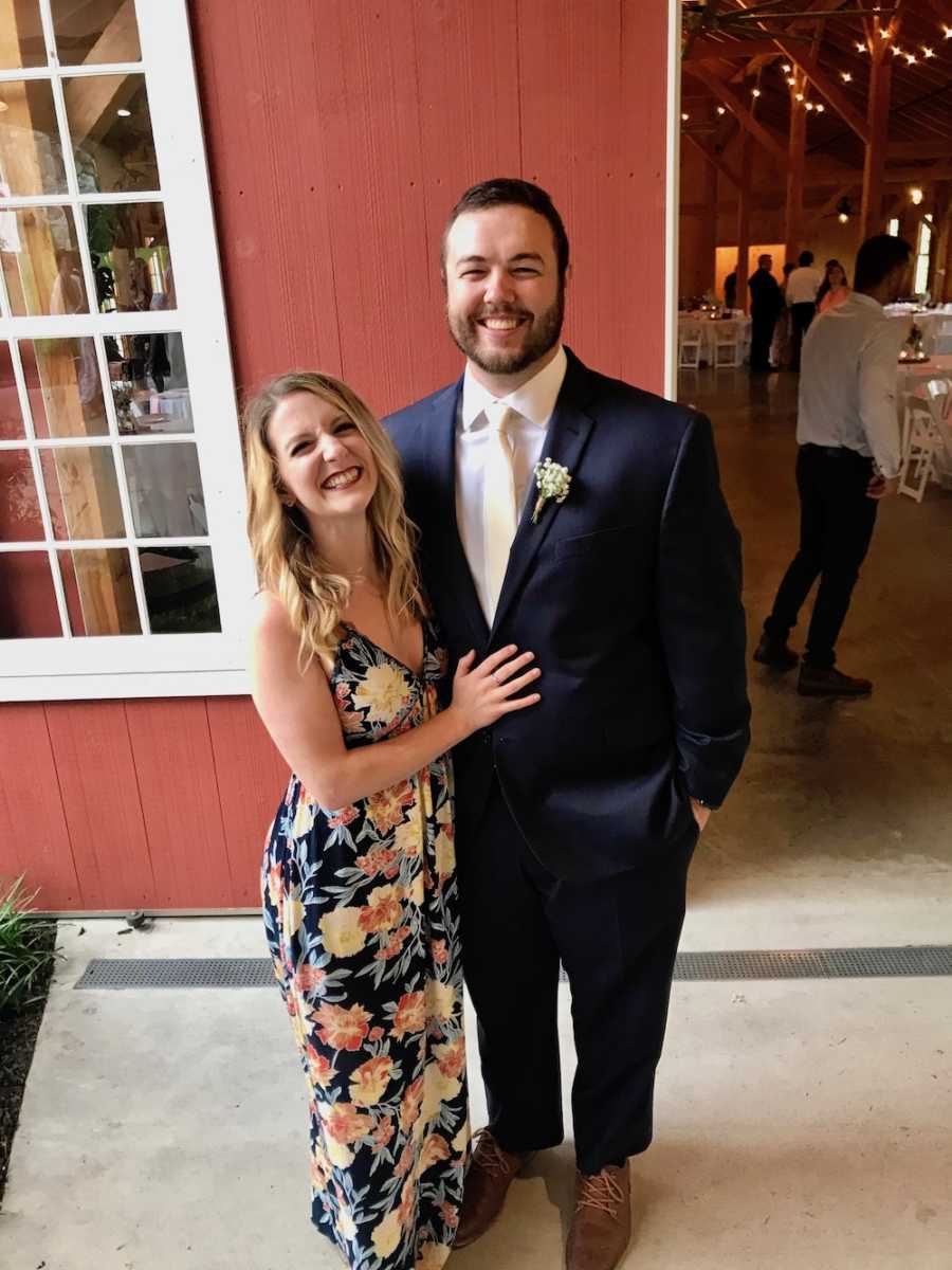 Husband and wife stand outside of red barn smiling in formal attire