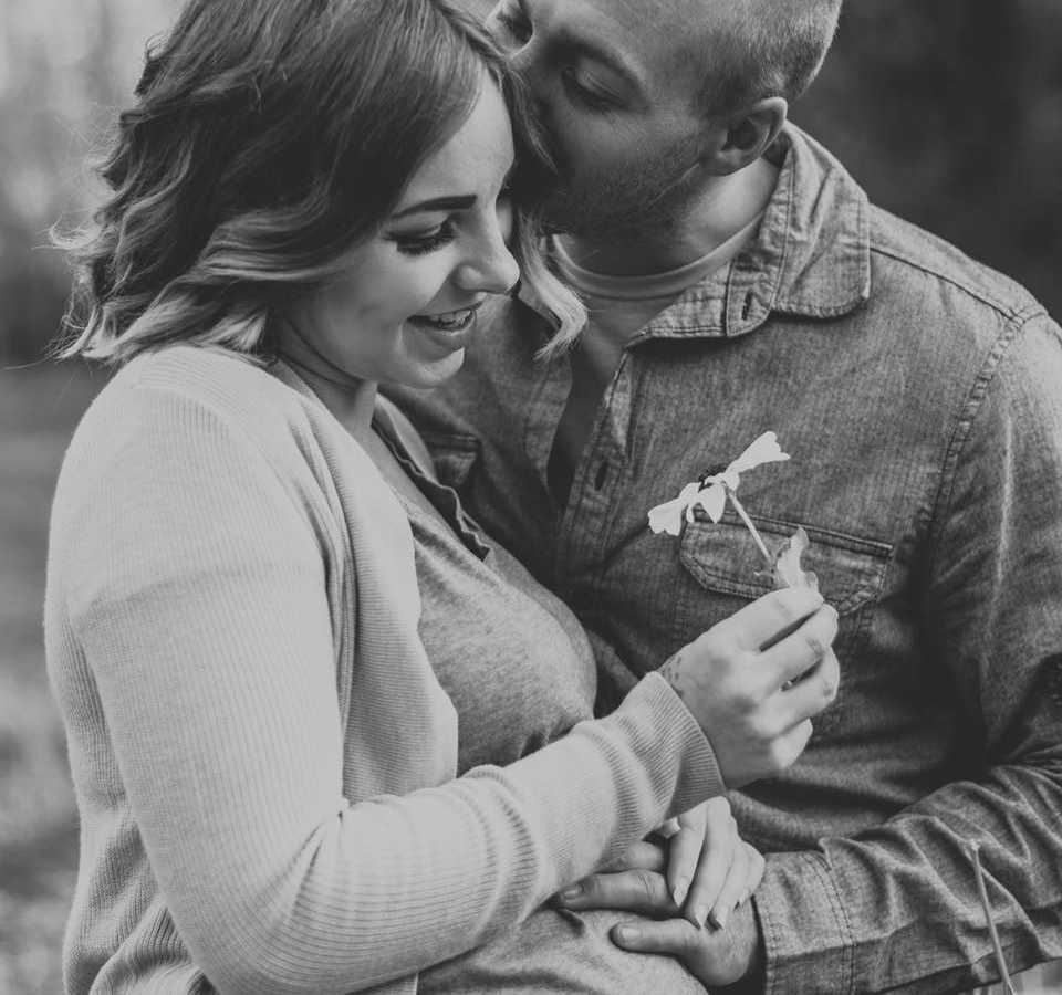 Pregnant woman smiles as she holds sunflower and husband kisses her head