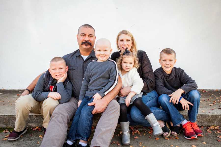 Husband and wife sit on curb with their four children, one of which has cancer