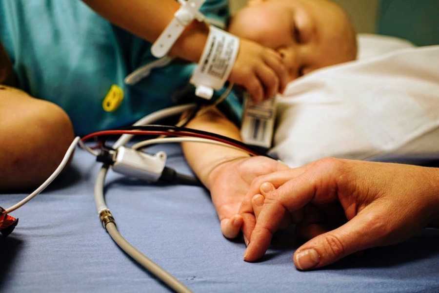 Little boy with cancer lays in hospital bed holding parent's hand