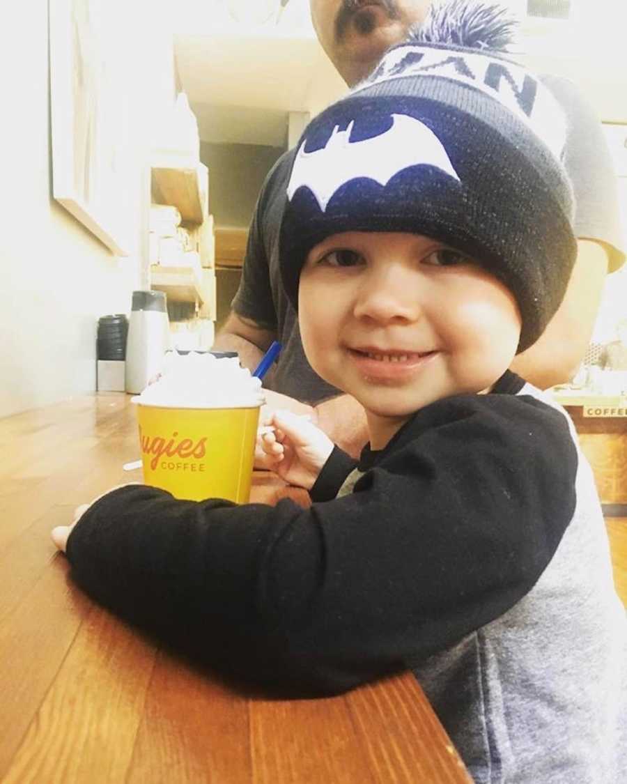 Little boy with cancer sits smiling at counter with drink with whip cream 