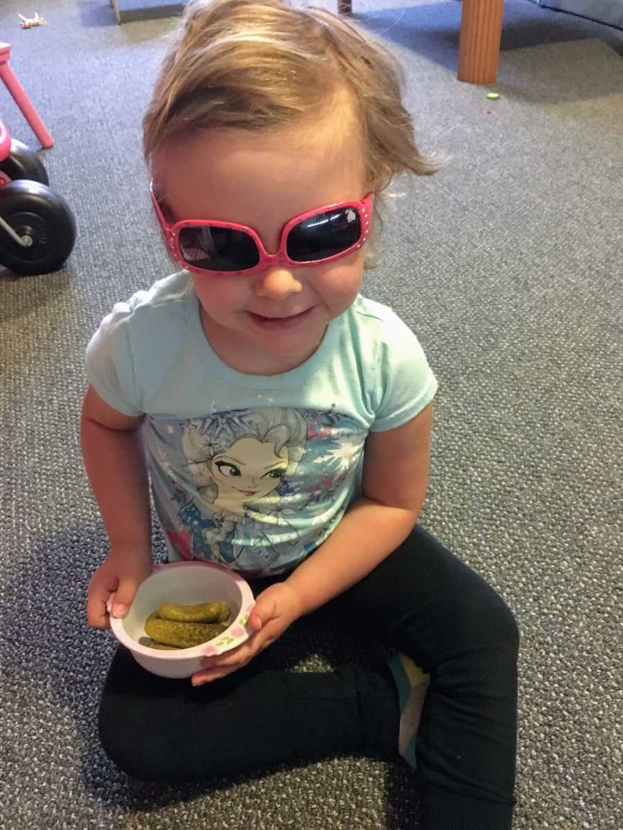 Little girl going through chemo sits on floor holding bowl of pickles while wearing pink sunglasses