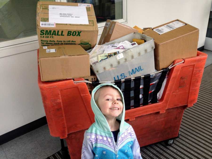 Little girl with cancer stands smiling in front of bin with boxes of post cards for her