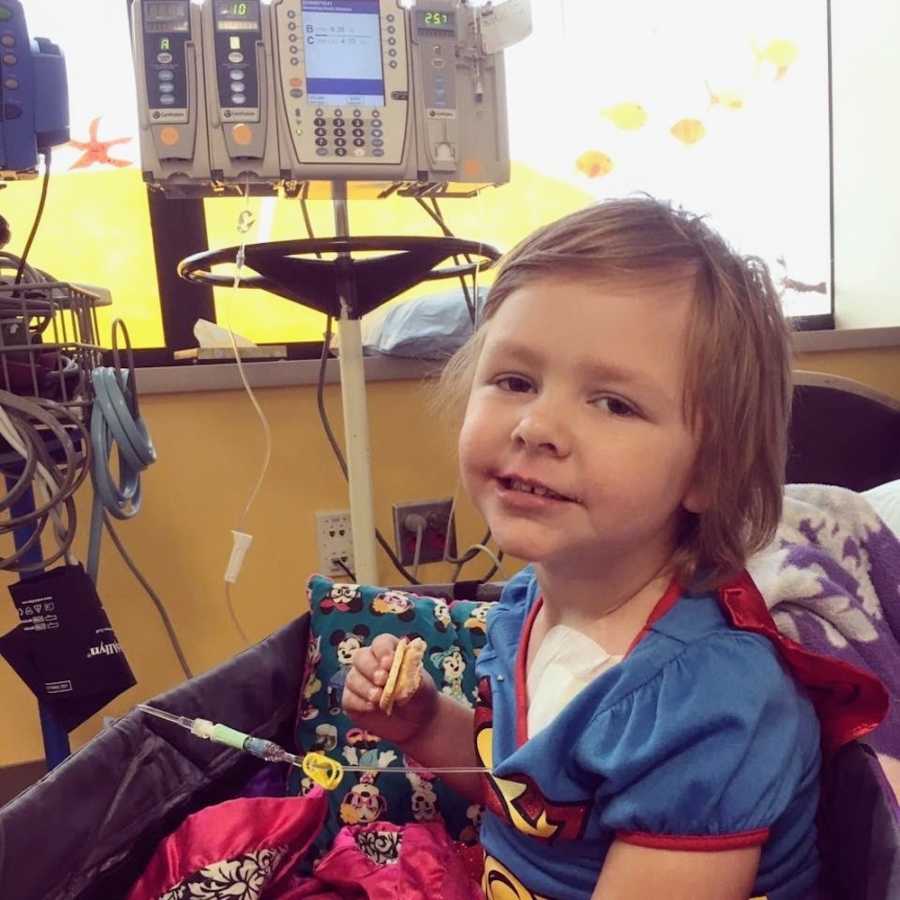 Little girl going through chemo sits in hospital bed smiles as she holds cheese and crackers
