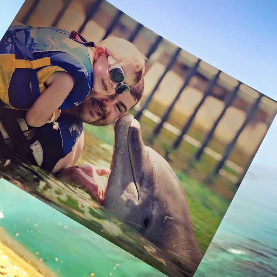 Little girl going through chemo smiles in pool with father as dolphin kisses his cheek