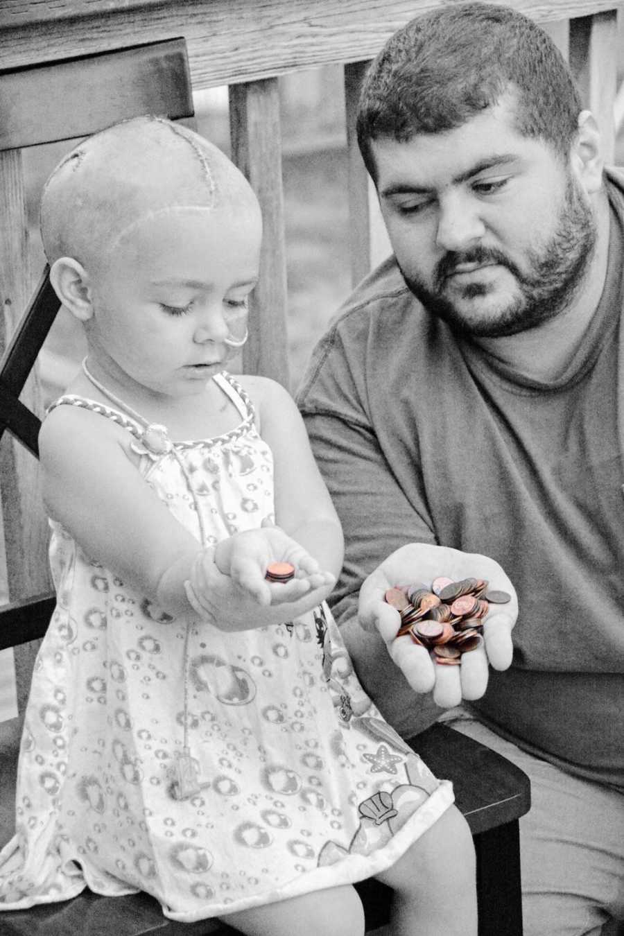 Little girl who has since passed away sits beside father as they hold out hand with pennies in them