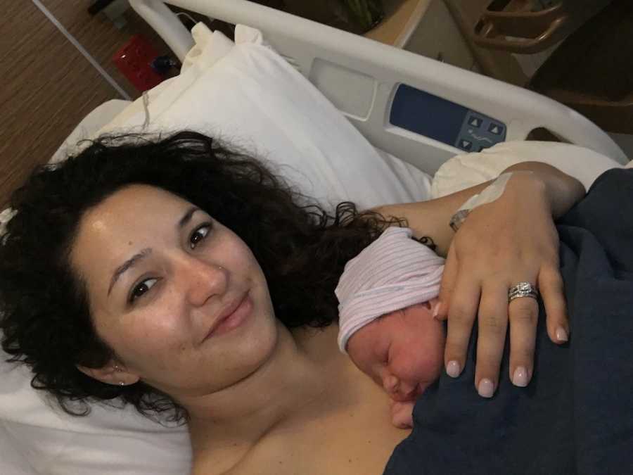 Woman smiles in selfie as she lies in hospital bed with newborn laying asleep on her bare chest