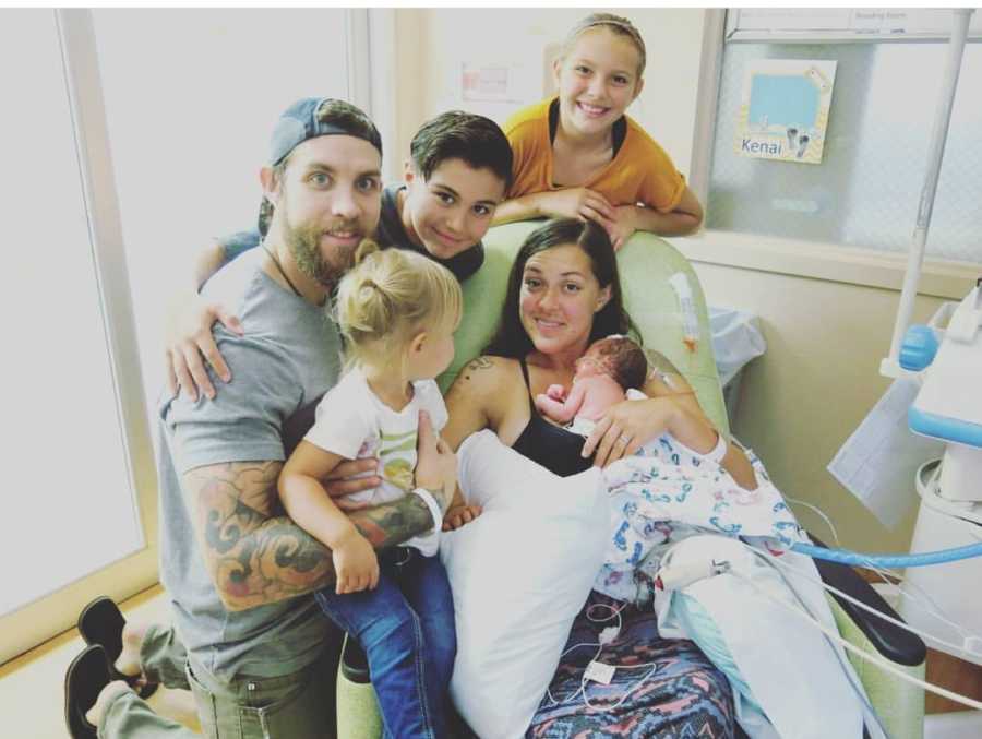 Woman sits in chair in NICU holding her newborn beside her husband and three other children