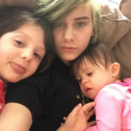 Oldest sibling sits on couch taking selfie with two younger siblings