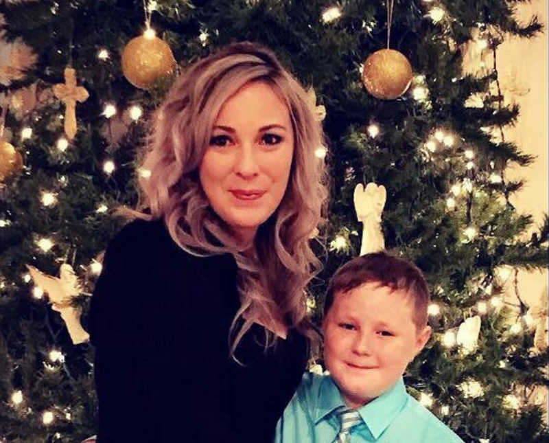 Mother who escaped abusive husband stands smiling in front of Christmas tree with young son 