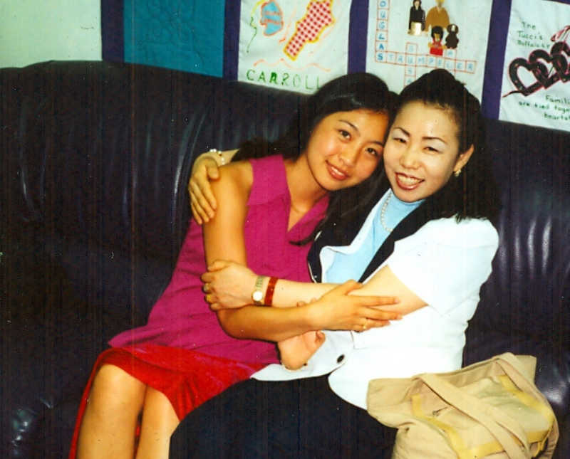 Young Korean woman sits on couch hugging biological mother who gave her up for adoption