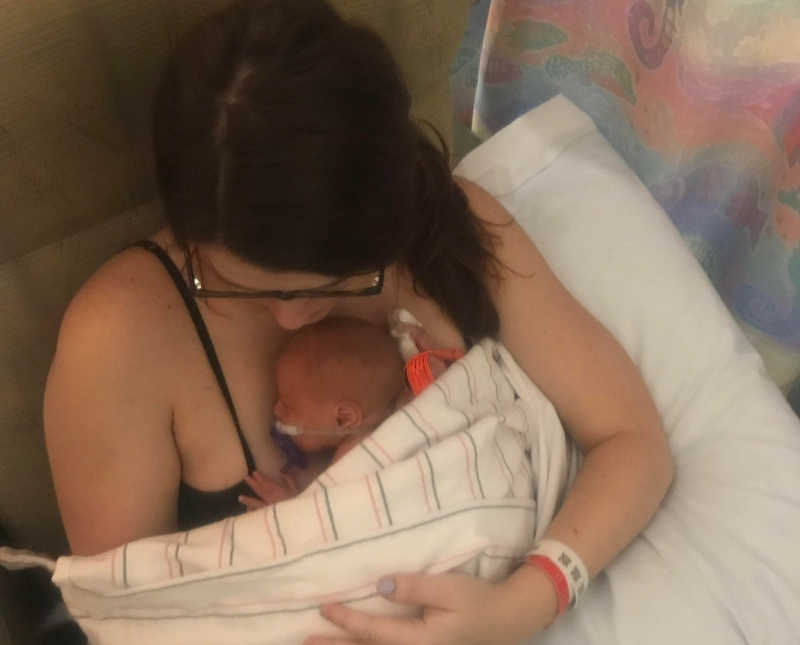 Woman sits in chair while newborn lays sleep on her chest with blanket over her