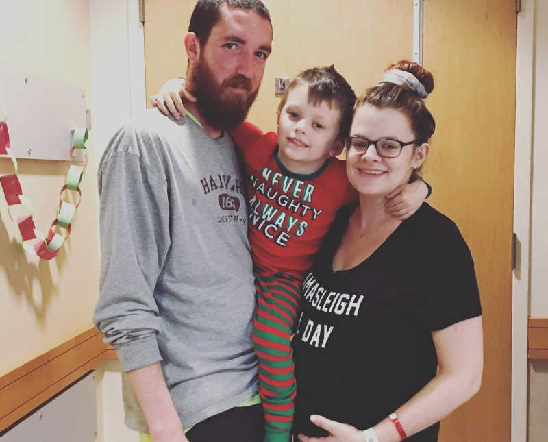 Pregnant woman stands in hospital room while holding her young son with her husband