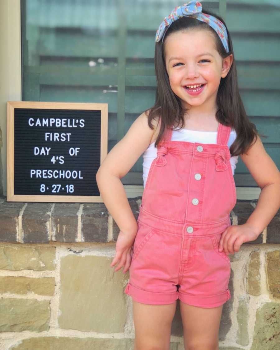 Little girl with autism stands outside beside sign announcing her first day of preschool