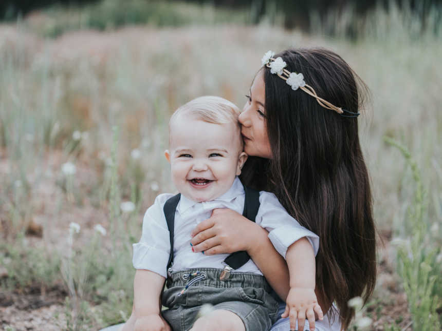 Little girl sits on ground in field outside kissing baby brother who sits on her lap