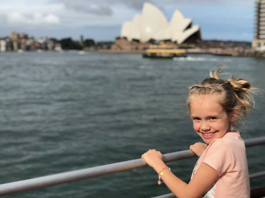 Little girl smiles as she holds onto rail beside body of water with Sydney Opera House in background