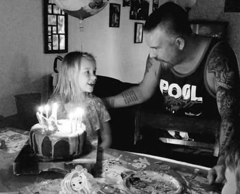 Little girl sits at table with birthday cake in front of her as she looks over smiling at mother's boyfriend