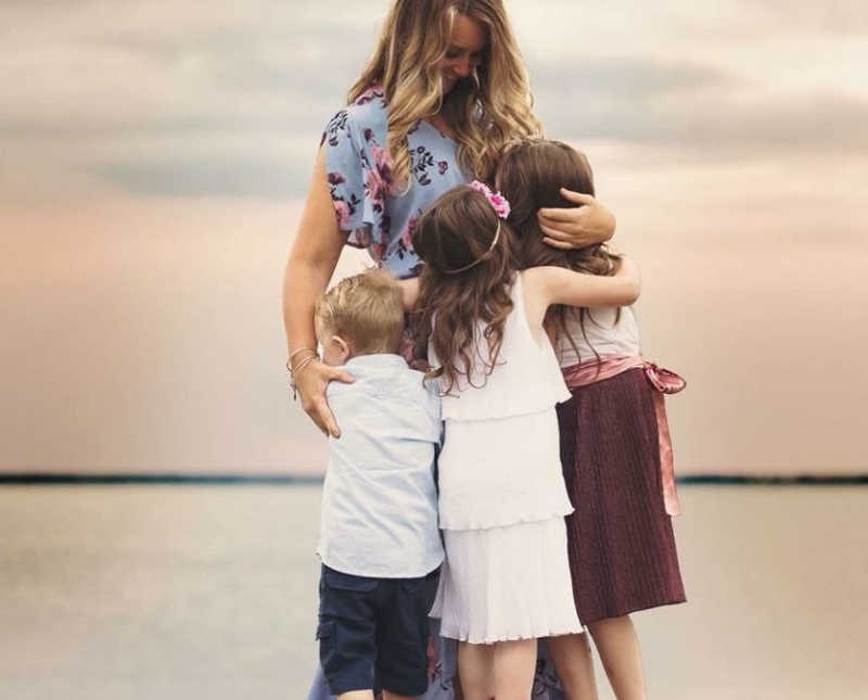 Widow stands smiling as her three kids hug her