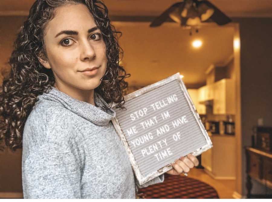 Woman holds sign in home that says, "Stop telling me that I'm young and have plenty of time"