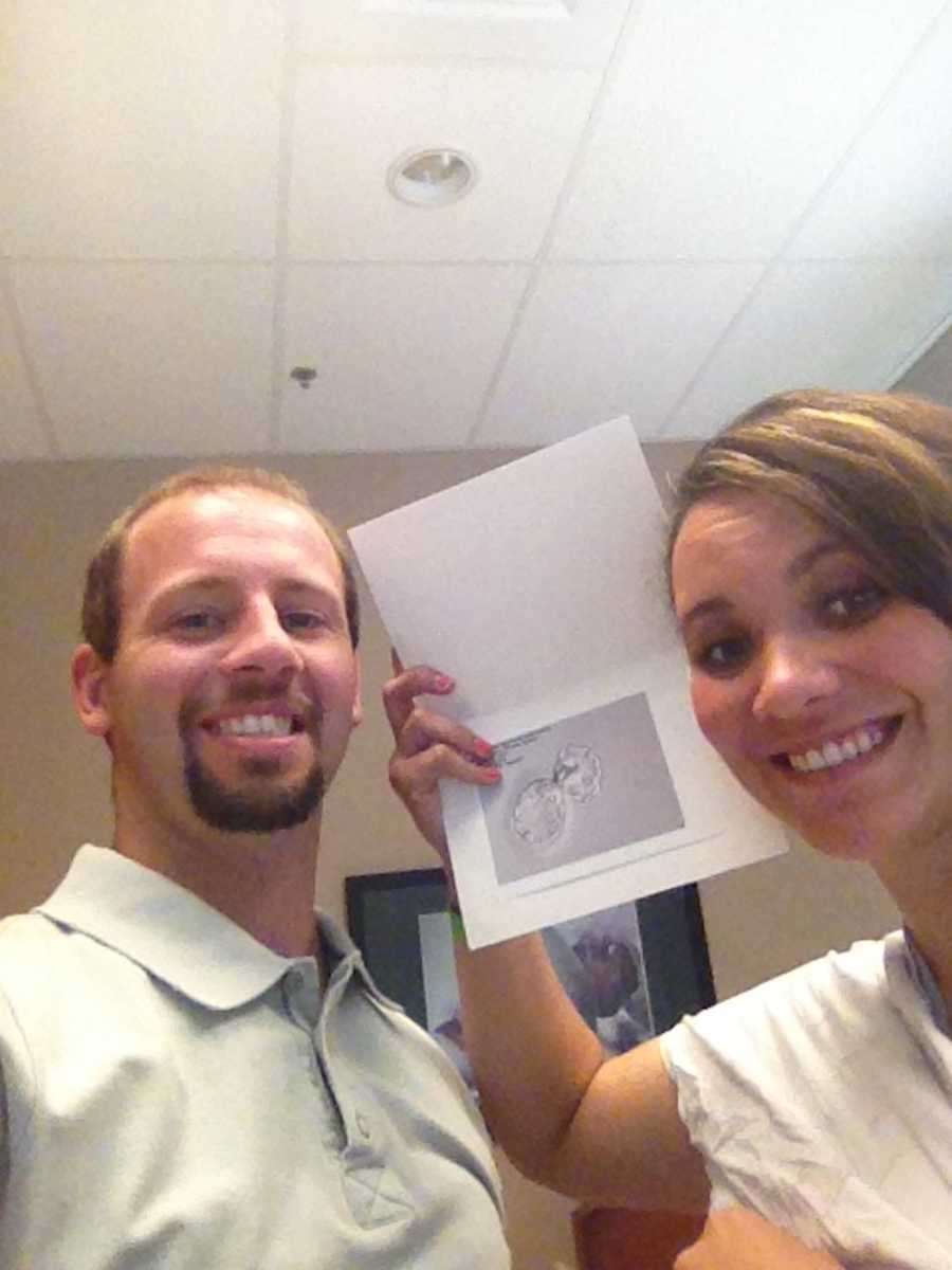Husband smiles beside wife whole holds up paper with ultrasound of implanted embryos