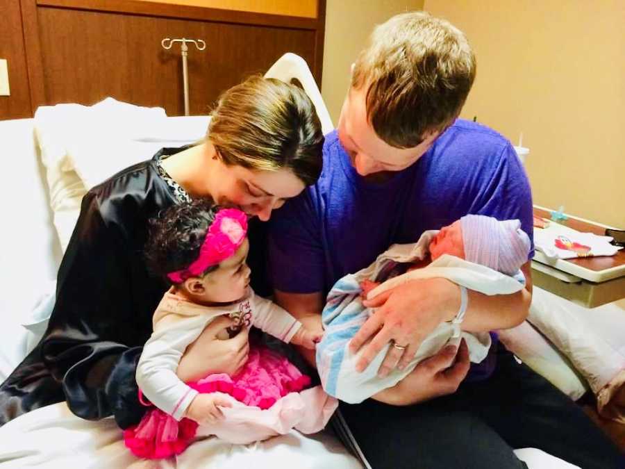 Woman sits up on hospital bed with adopted daughter beside husband and newborn