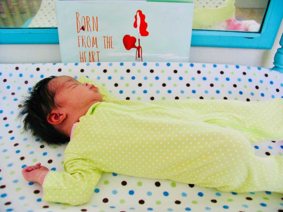 Adopted newborn lays asleep on her back beside sign that says, "Born from the heart"