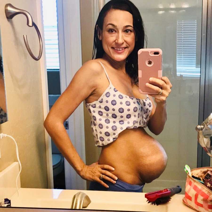 Pregnant woman smiles in mirrors selfie in bathroom with shirt lifted above her stomach