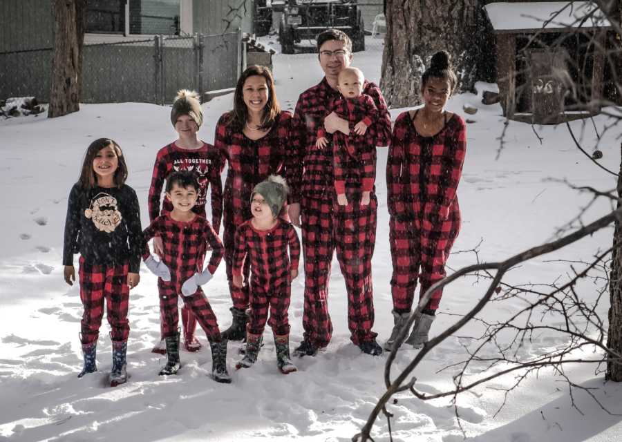 Husband and wife stand outside in snowy weather with their newborn beside their five foster children