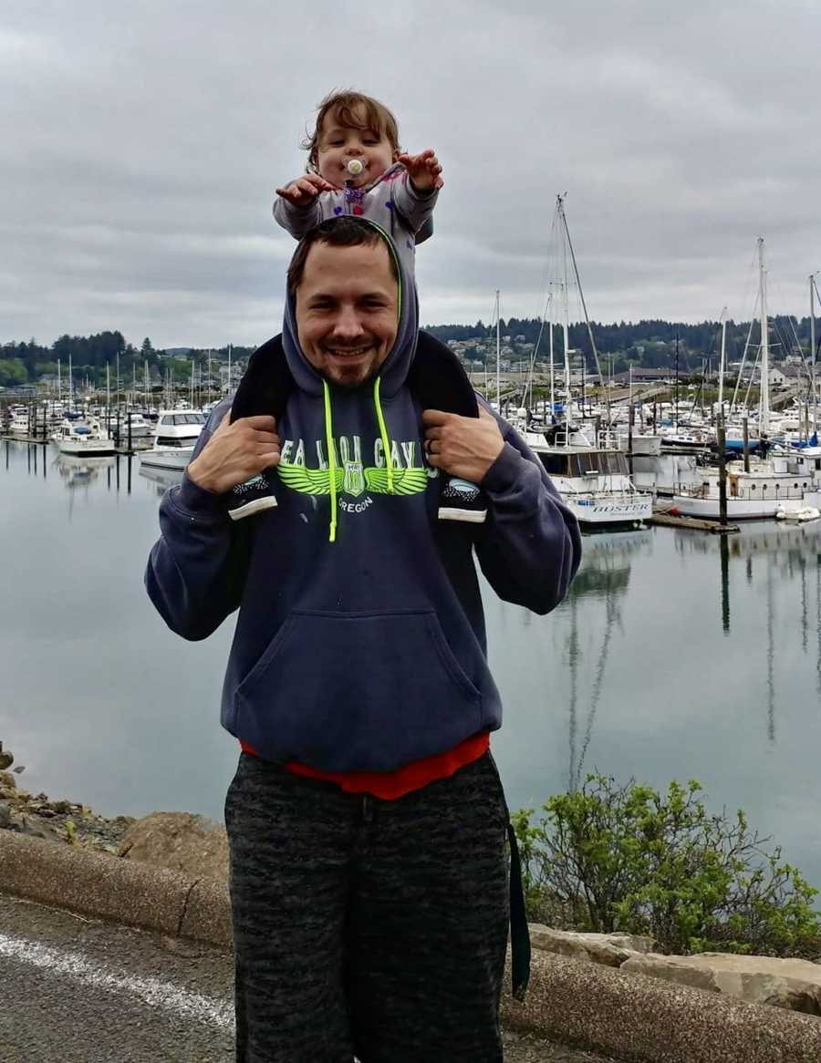 Man stands with young daughter on his shoulders beside marina