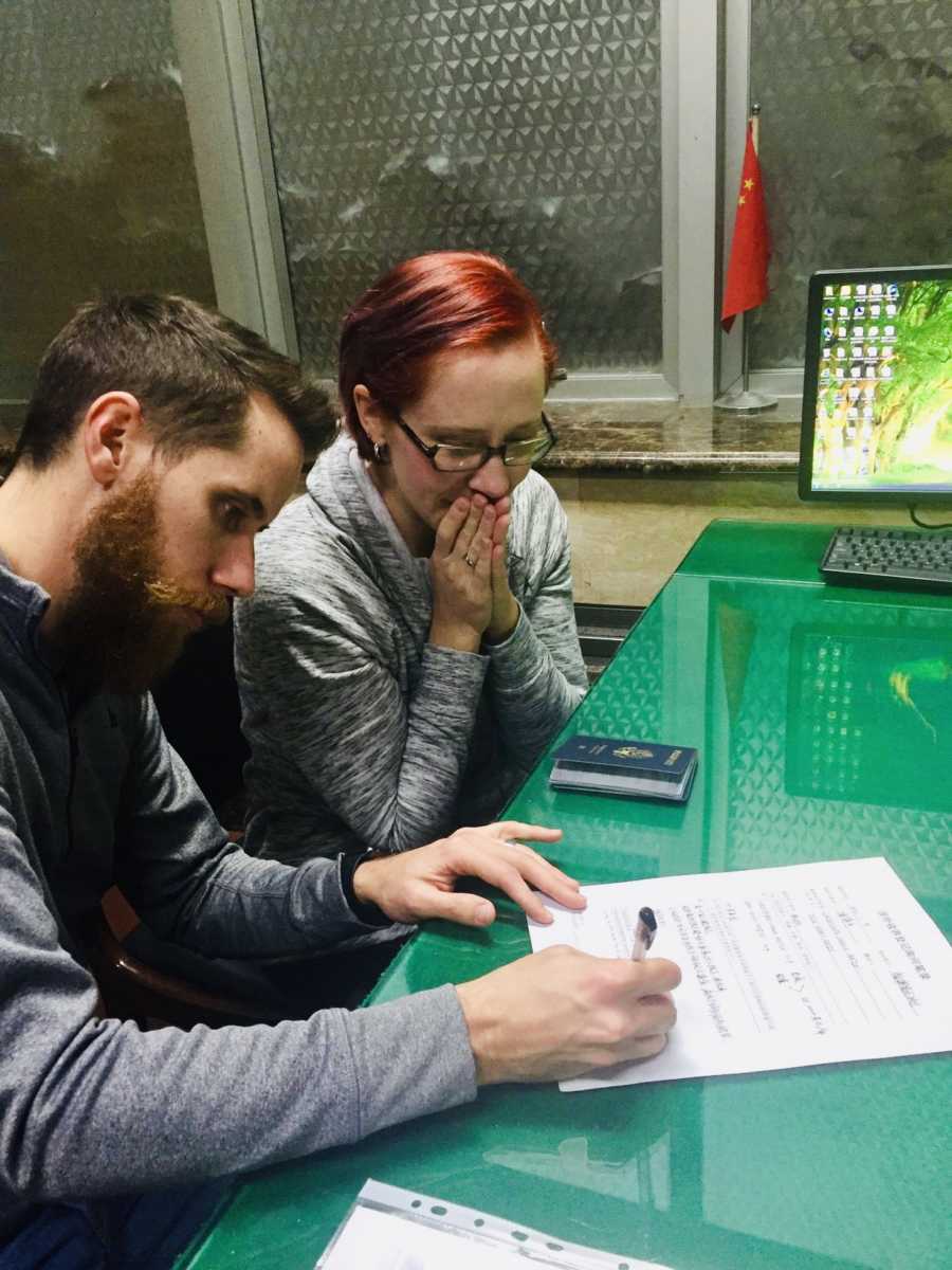 Husband signs adoption paperwork while wife holds hands to mouth watching