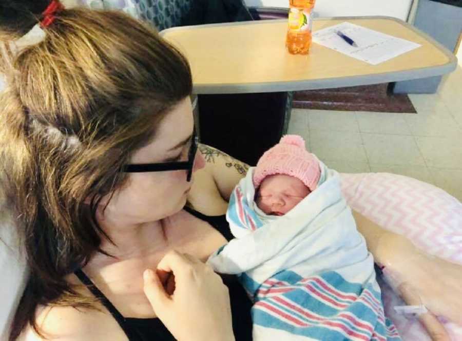 Woman sits in hospital room with newborn girl asleep in her lap swaddled in blanket