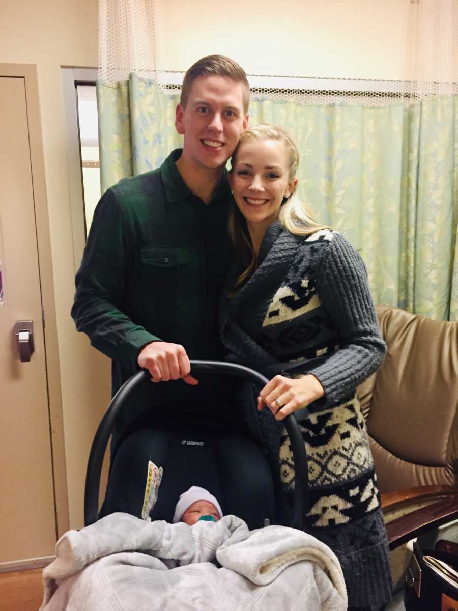 Husband and wife stand in hospital room smiling as they hold carseat where adopted son sleeps