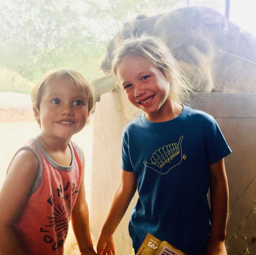 Brother and sister who beat brain cancer stand smiling at zoo with zoo animal in background