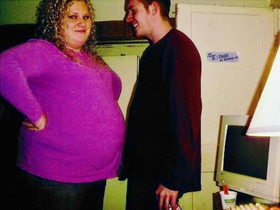 Pregnant woman stands with hands on hips smiling as her husband looks at her smiling