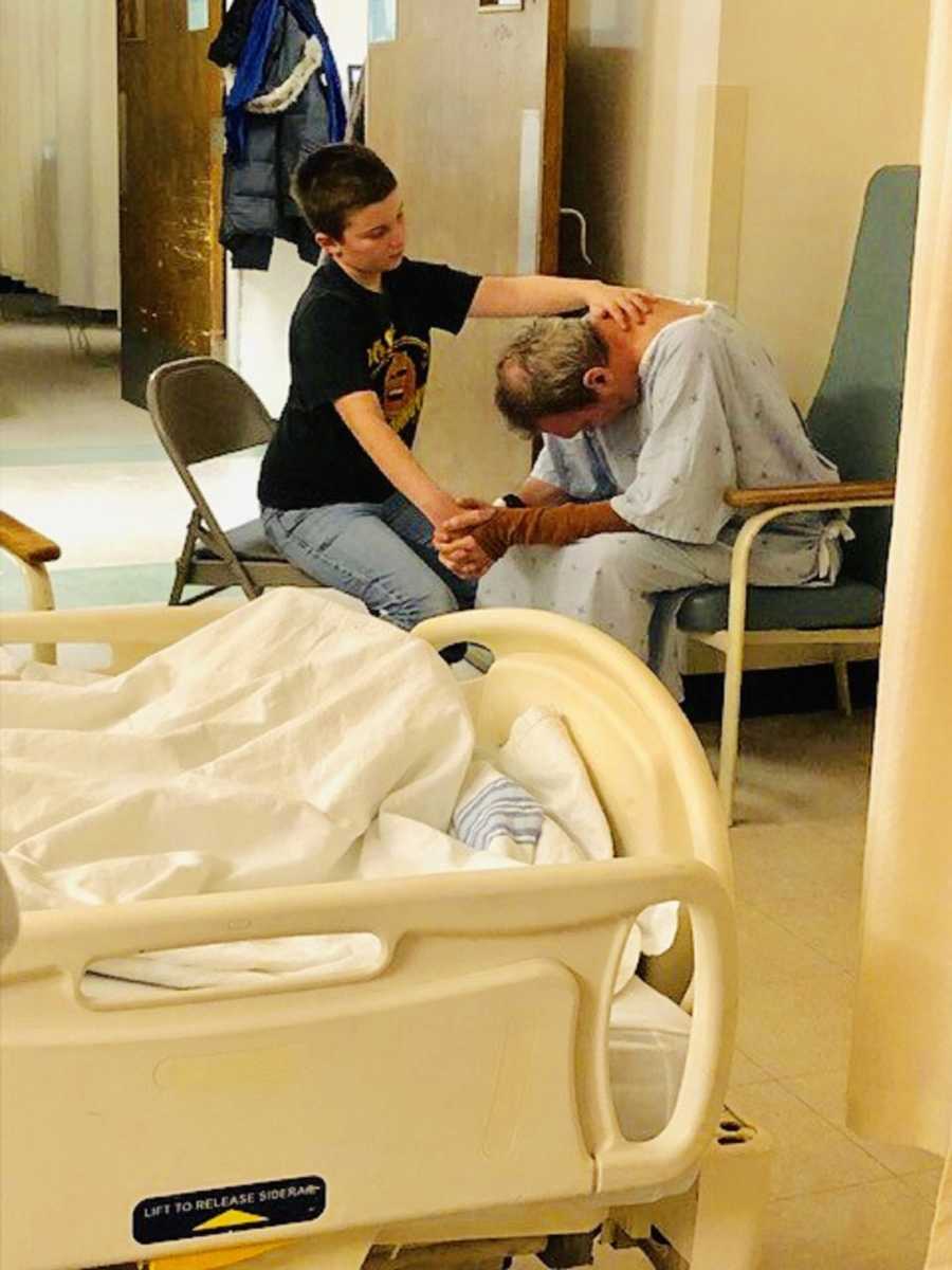 Man who had brain tumor and seizure sits leaned over in chair in hospital room holding grandson's hand