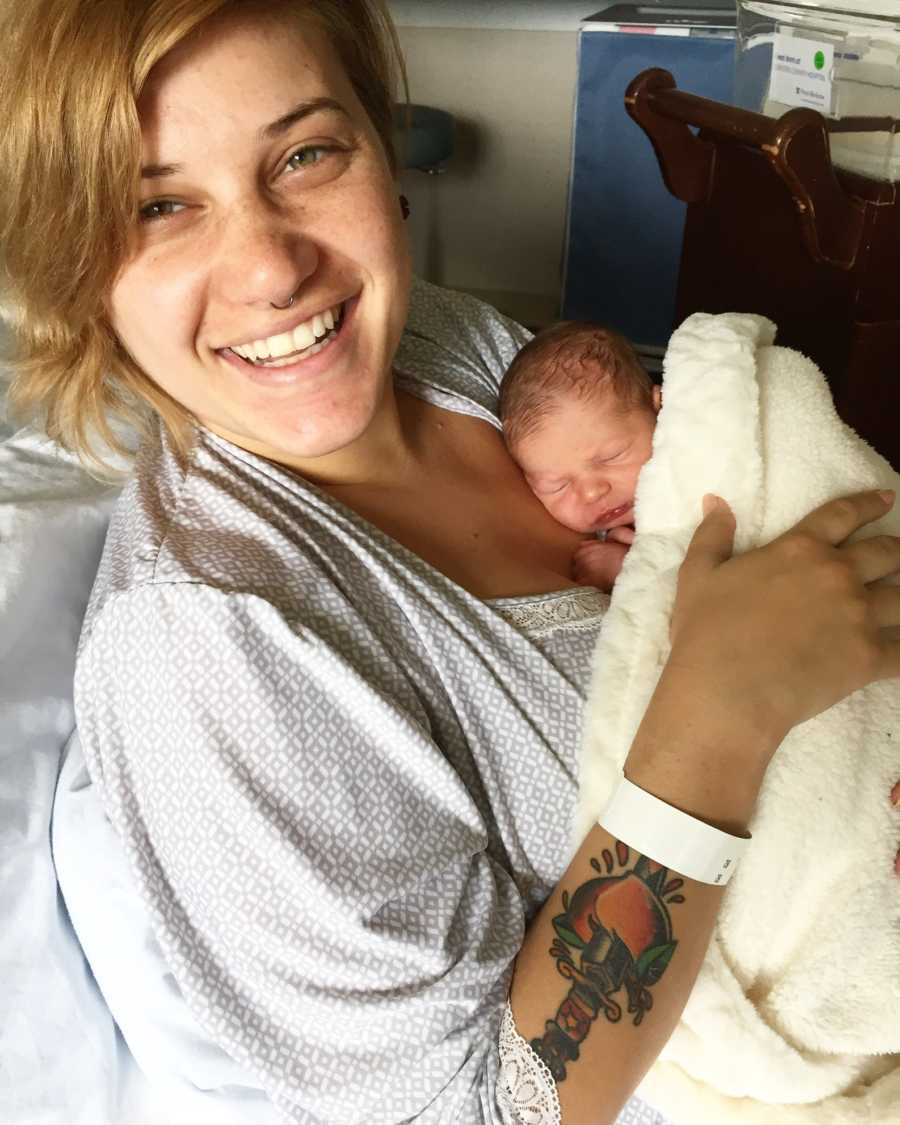 Woman smiles while lying in hospital bed with newborn asleep on her chest