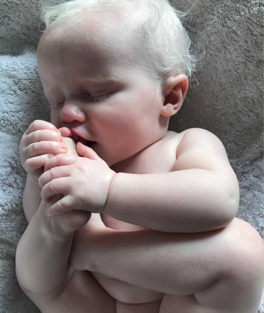 Albino baby lays on gray blanket holding foot up to mouth