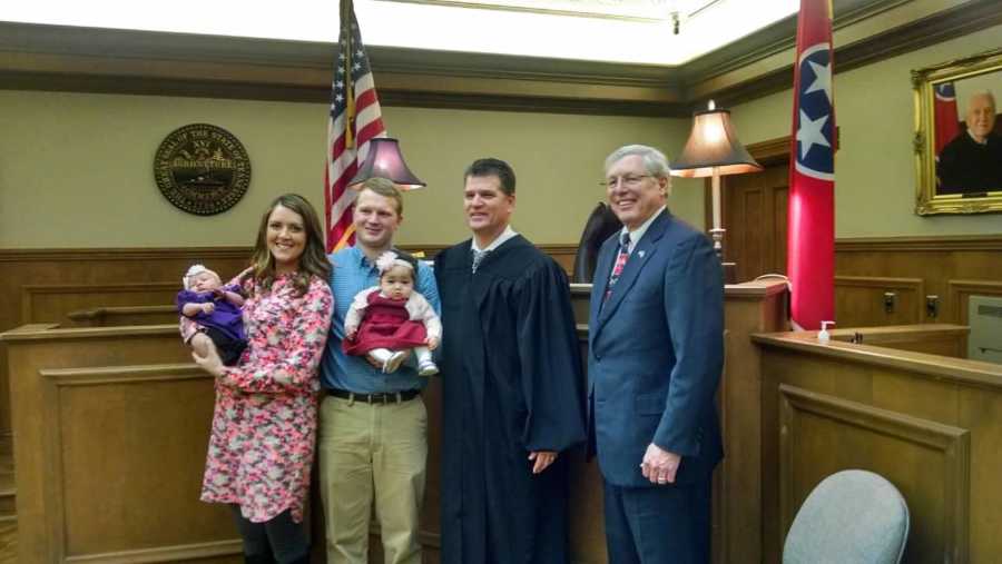 Husband and wife stand in adoption court with their two baby girl's beside judge and another man