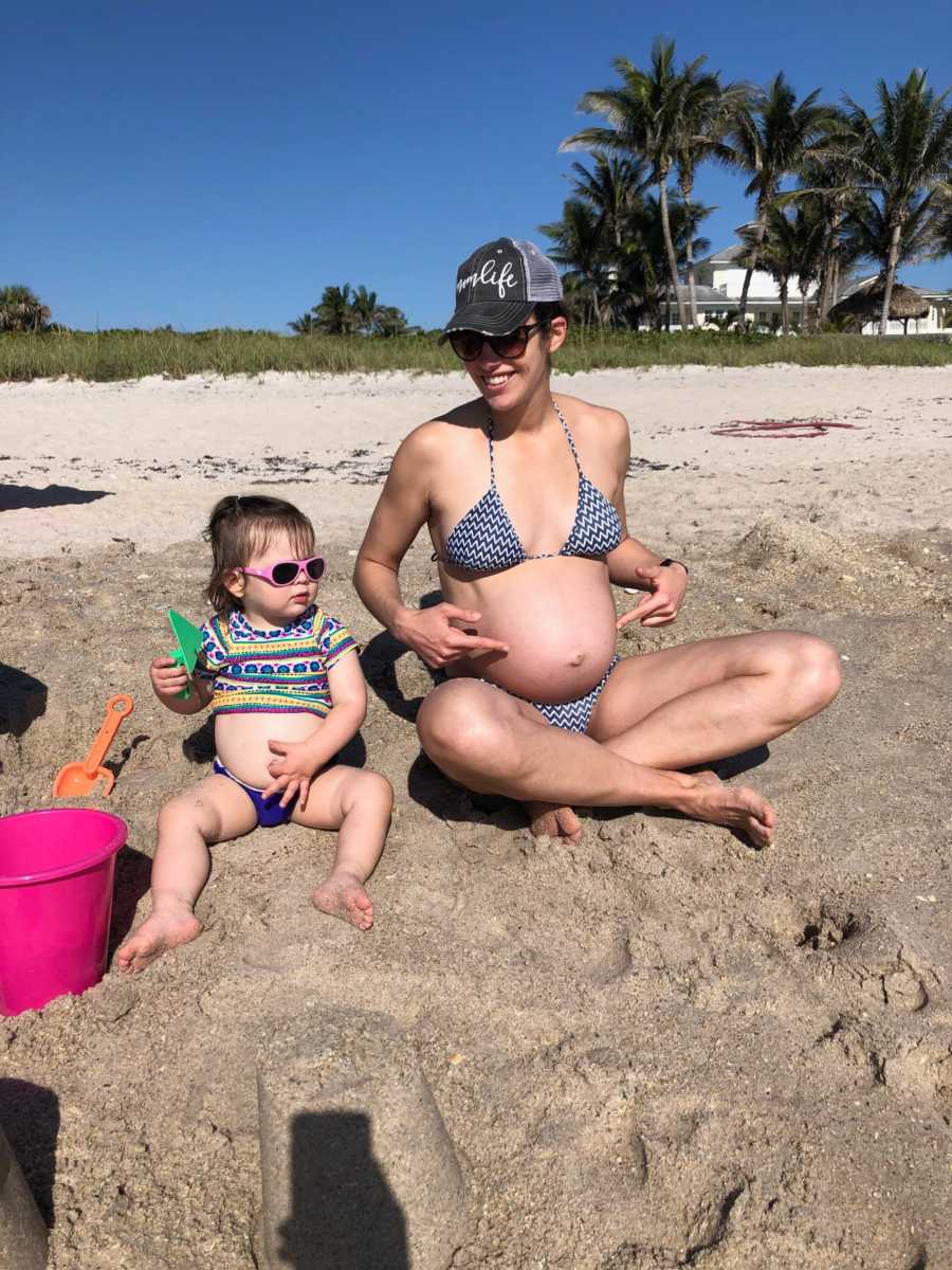 Pregnant woman sits on beach in bikini beside daughter