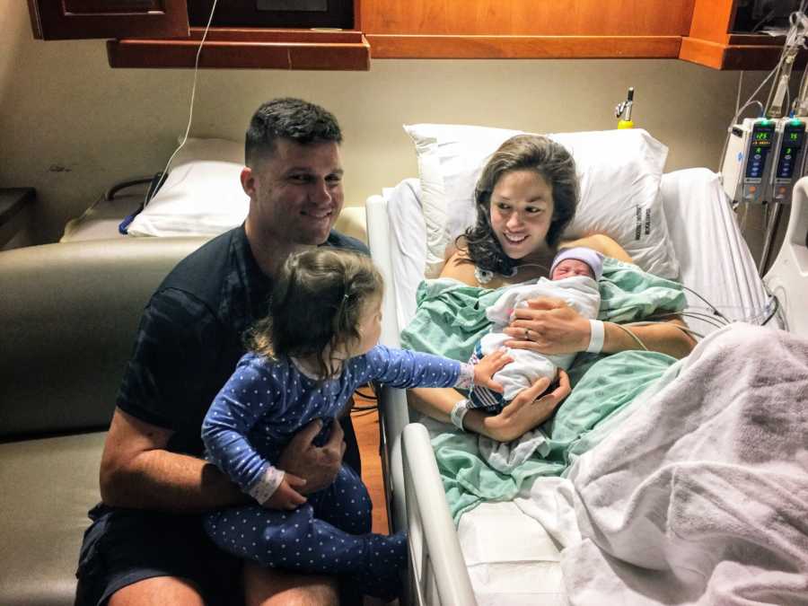 Mother smiles in hospital bed holding newborn as husband and first born sit beside them