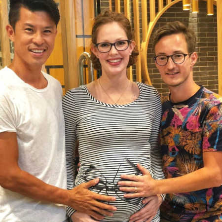 Pregnant gestational carrier stands smiling while gay couple stand on either side of her holding her stomach