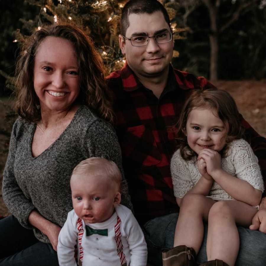 Mother and baby son with Ichthyosis sit outside beside father and daughter in front of tree with lights on it