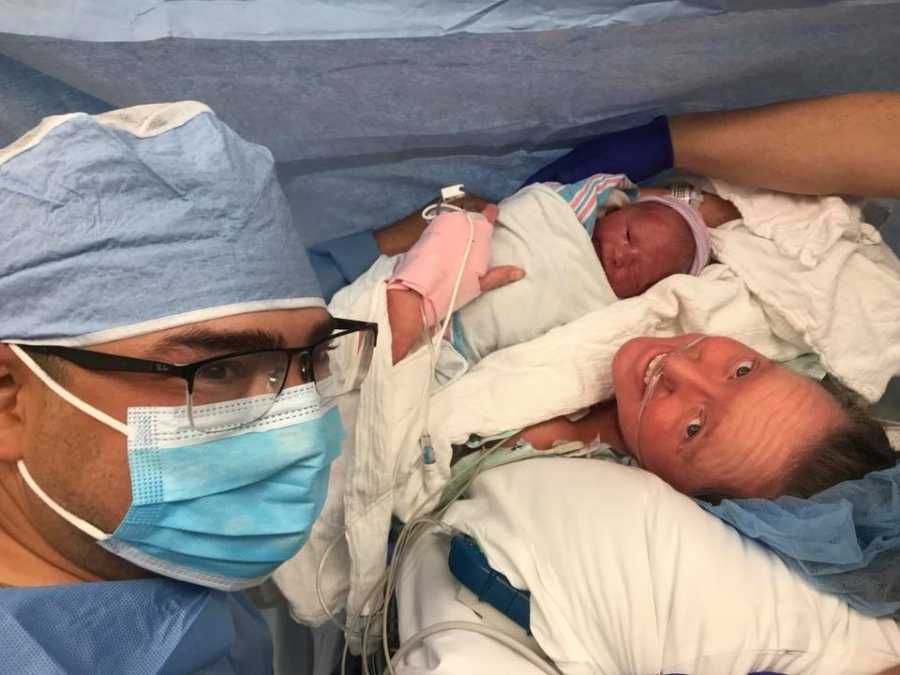 Father in scrubs and mask takes selfie with wife who just had c-section who holds newborn