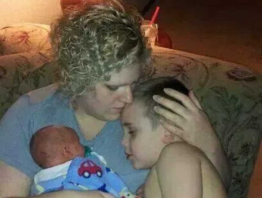 Mother sits with son resting head on her shoulder while she holds newborn 