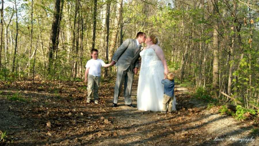 Bride and groom stand kissing in path of woods while holding hands with their two sons