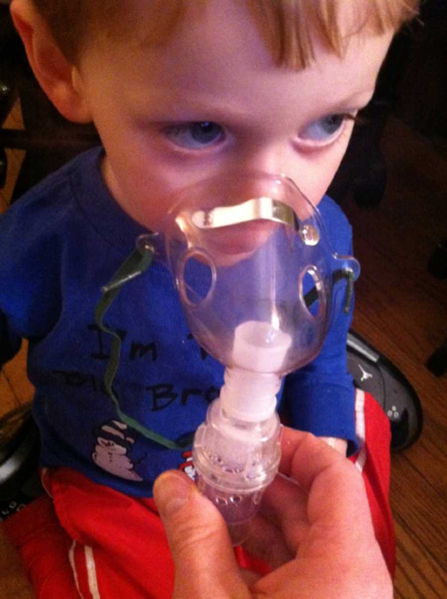 Little boy sits on floor while adult holds oxygen mask up to his mouth