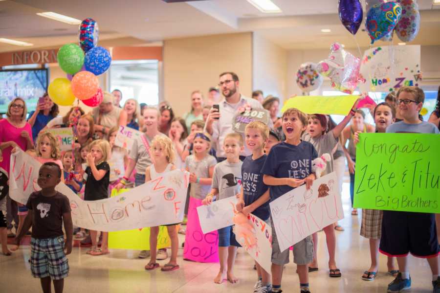 Crowd of kids and adults holding signs and balloons at airport for arrival of adopted girl