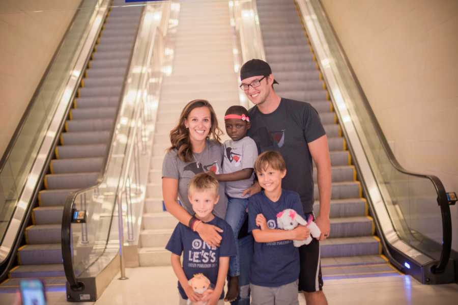 Husband and wife stand by escalators at airport holding adopted daughter with their two sons