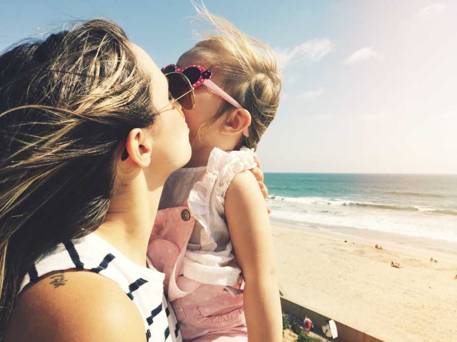 Mother holds and kisses daughter on cheek with beach in background